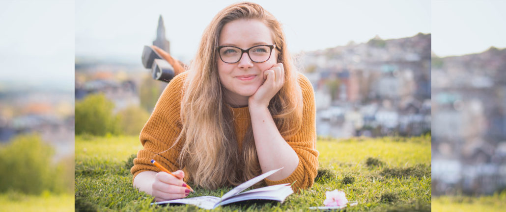 young woman laying on her belly on a field, journaling, with blurry cityscape in the background