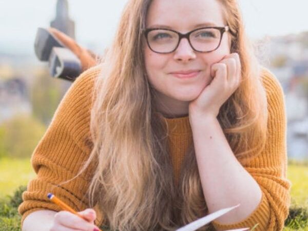 young woman laying on her belly on a field, journaling, with blurry cityscape in the background