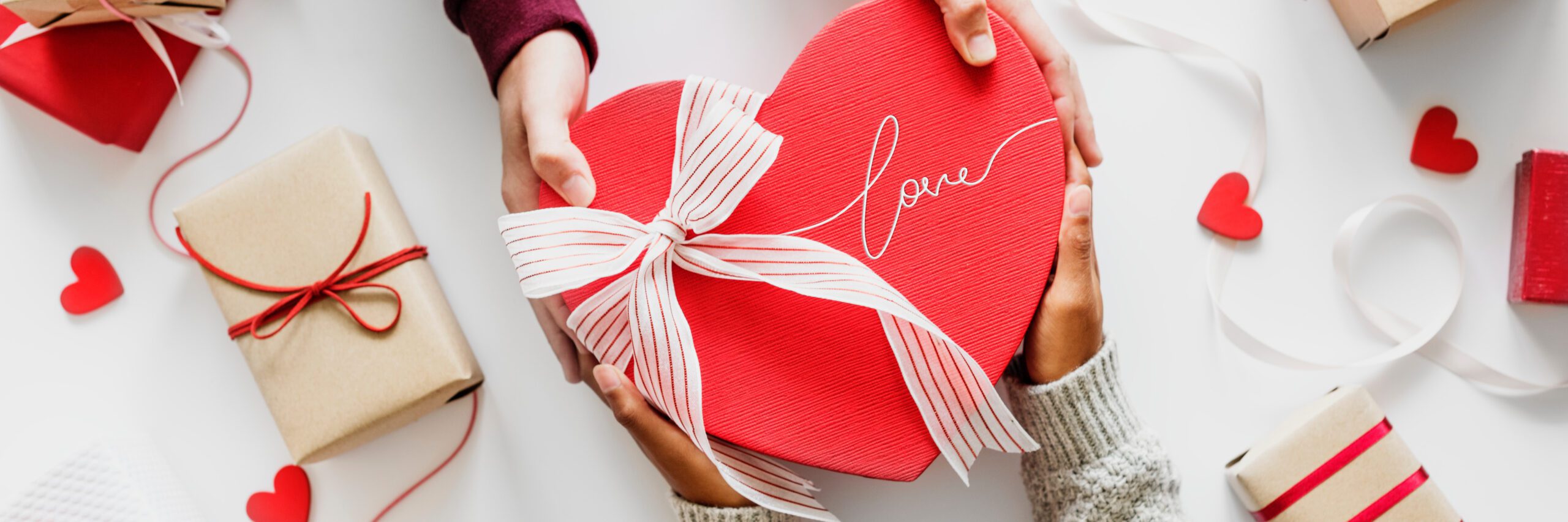 two sets of hand holding a heart shaped box with "Love" engraved on it on a bow tied on it, surrounded by presents and paper hearts