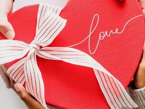 two sets of hand holding a heart shaped box with "Love" engraved on it on a bow tied on it, surrounded by presents and paper hearts