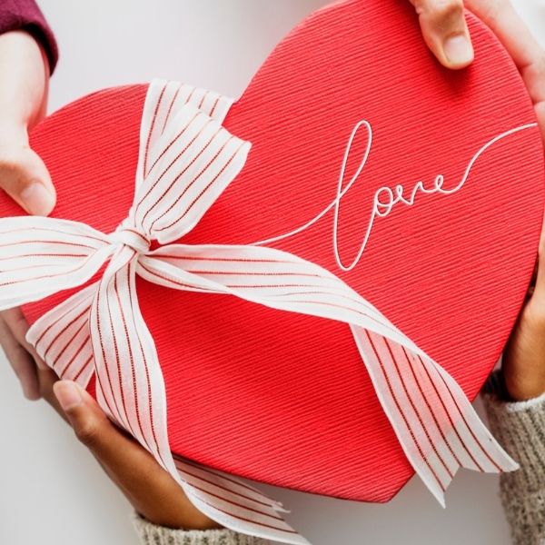 two sets of hand holding a heart shaped box with "Love" engraved on it on a bow tied on it, surrounded by presents and paper hearts