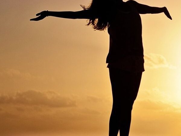 silhouette of a young woman with arms stretched out, with the ocean behind them at sunset