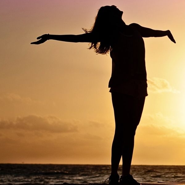 silhouette of a young woman with arms stretched out, with the ocean behind them at sunset