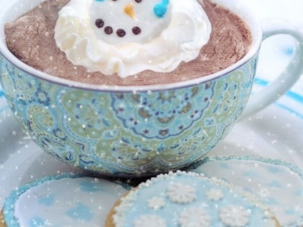 blue mug filled with hot chocolate topped with whip-cream with a snowman pipped on it, on a plate of cookies