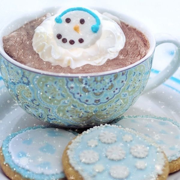 blue mug filled with hot chocolate topped with whip-cream with a snowman pipped on it, on a plate of cookies